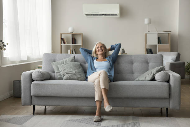 Cheerful old woman resting on grey soft couch under conditioner
